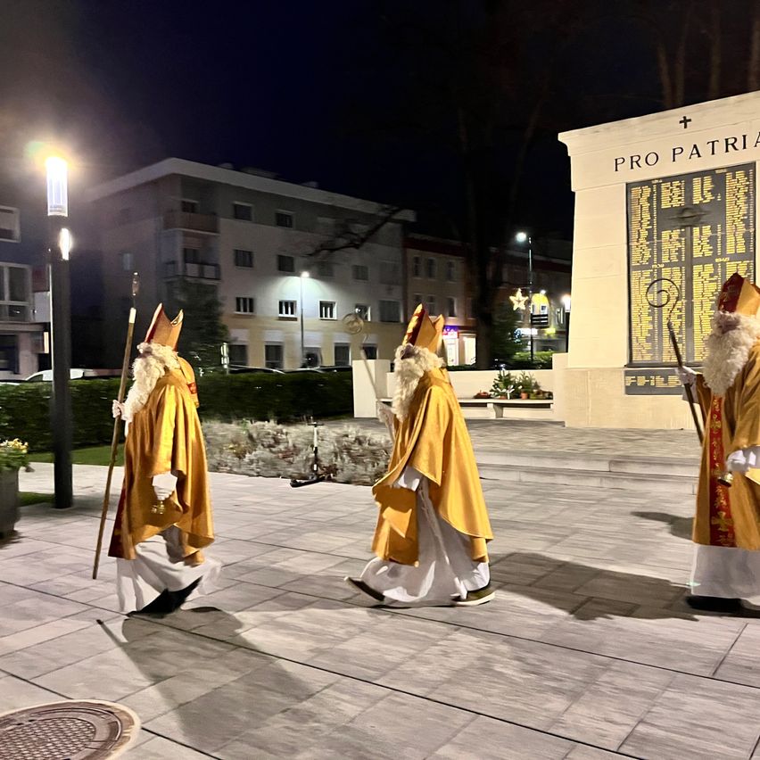 Drei Männer, gekleidet in gelbe Roben und Mützen, gehen nachts auf einem gepflasterten Weg. Sie halten Stöcke in den Händen und scheinen eine Zeremonie durchzuführen. Hinter ihnen steht ein Gebäude mit einer Wand, auf der die Worte 'PRO PATRIA' stehen. Es gibt Straßenlaternen, eine Bank und Pflanzen in der Umgebung.