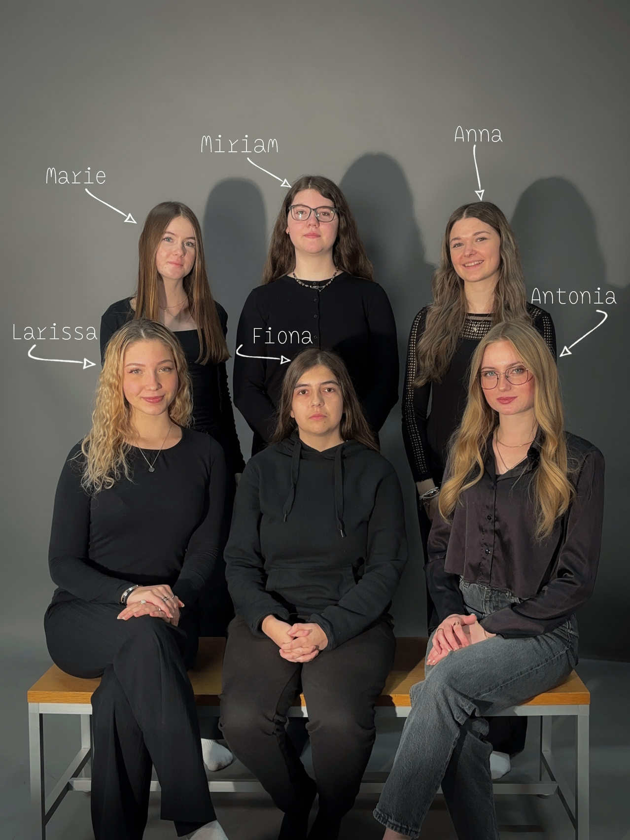 Eine Gruppe von Frauen sitzt auf einer Bank, jede mit einem Namen versehen: Miriam, Marie, Fiona, Larissa, Antonia und Anna. Sie tragen alle schwarze Kleidung und scheinen für ein Foto zu posieren.