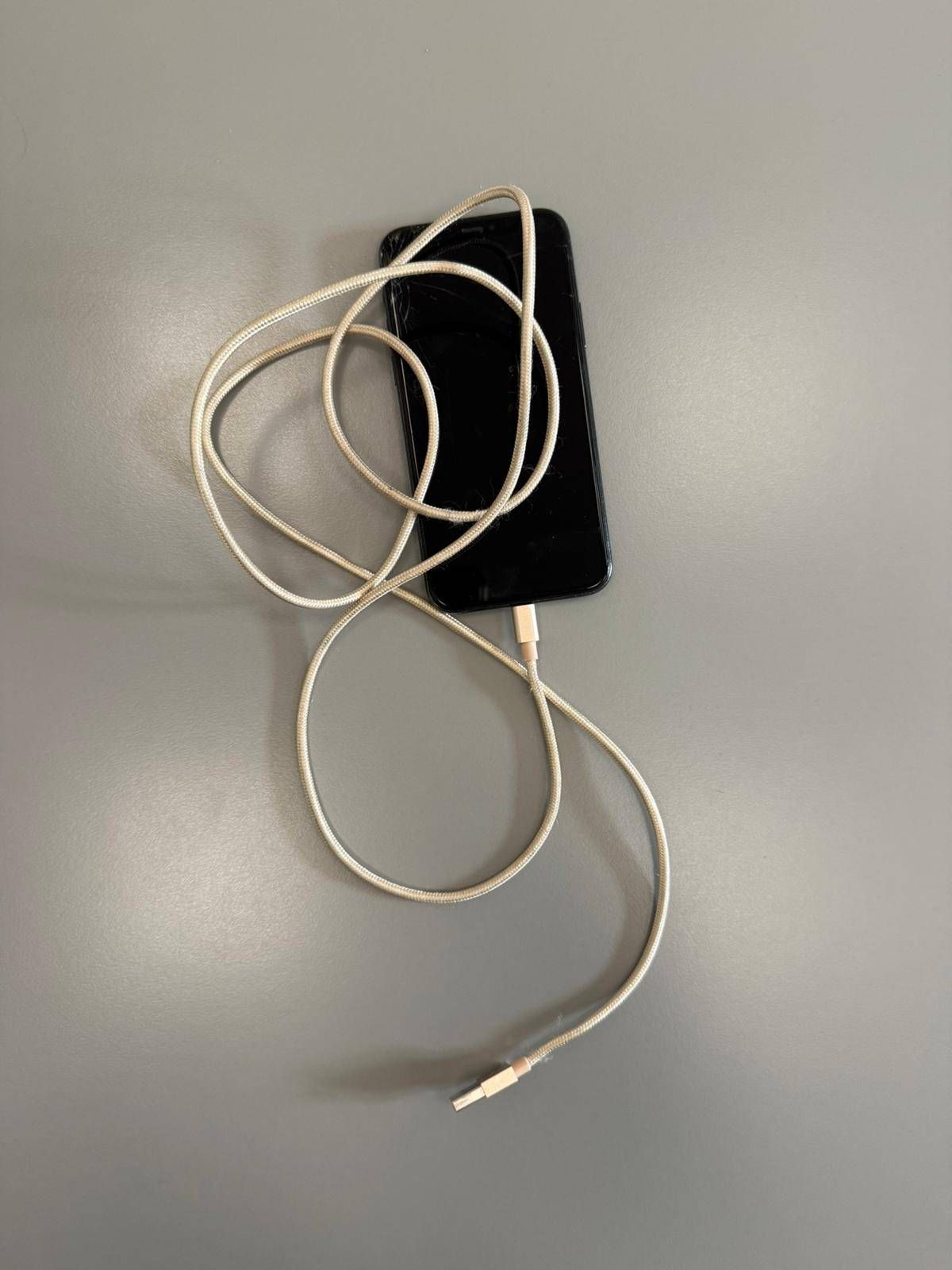 Ein schwarzes Smartphone, das mit einem weißen Ladekabel mit goldfarbenem Stecker verbunden ist, liegt auf einer grauen Oberfläche. Das Kabel ist aufgewickelt und scheint in Gebrauch zu sein.