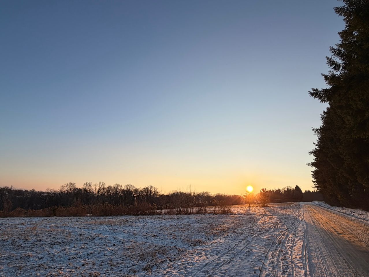 Ein Winter-Sonnenuntergang über einer verschneiten Landschaft mit klarem Himmel, Bäumen und einem kleinen Feld mit Reifenspuren.