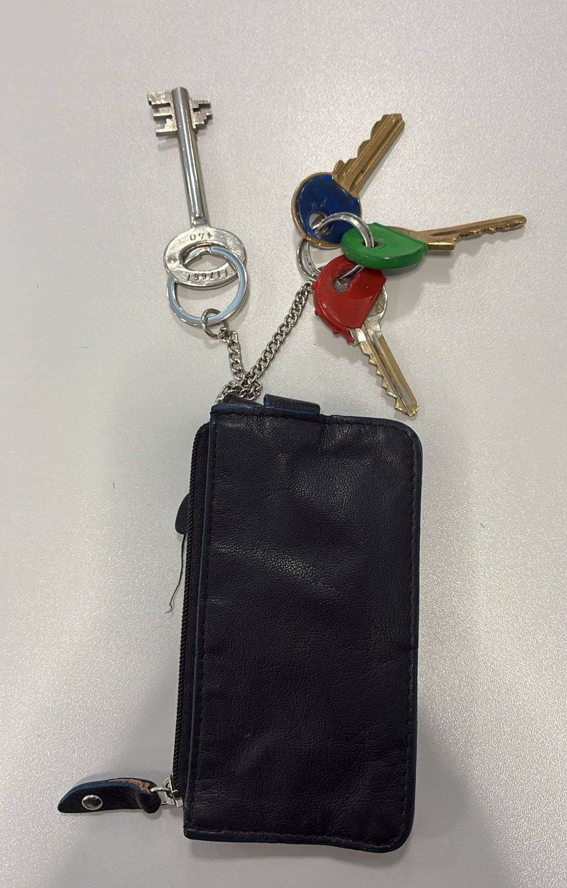 Ein schwarzer Ledergeldbeutel mit einem silbernen Schlüsselbund, der einen Hauptschlüssel und drei farbige Schlüssel enthält. Die Schlüssel sind mit einem silbernen Ring und einer silbernen Kette verbunden.