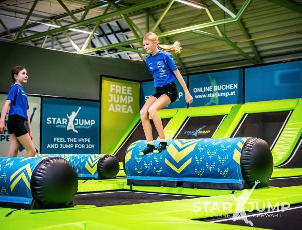 Zwei Mädchen springen auf einem Trampolin mit Star Jump Werbung. Das Mädchen in Blau ist in der Luft, während das Mädchen in Blau hinter ihr geht.