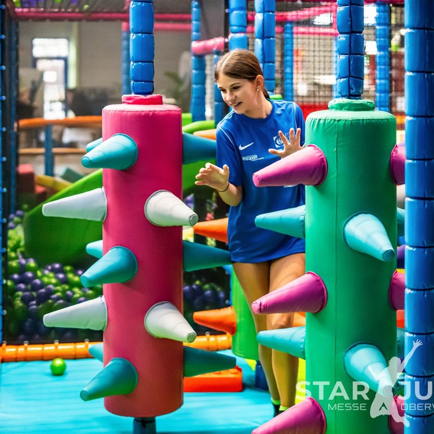 Eine junge Frau in einem blauen Shirt navigiert durch einen farbenfrohen aufblasbaren Spielbereich, umgeben von Kegeln und bunten Hindernissen.