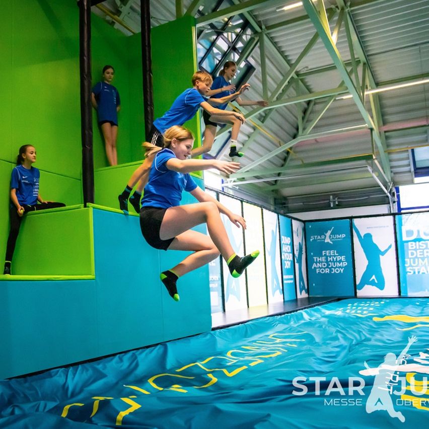 Eine Gruppe junger Menschen in blauen Hemden und Shorts springt auf einem Trampolin in einer Turnhalle mit grünen und blauen Wänden.
