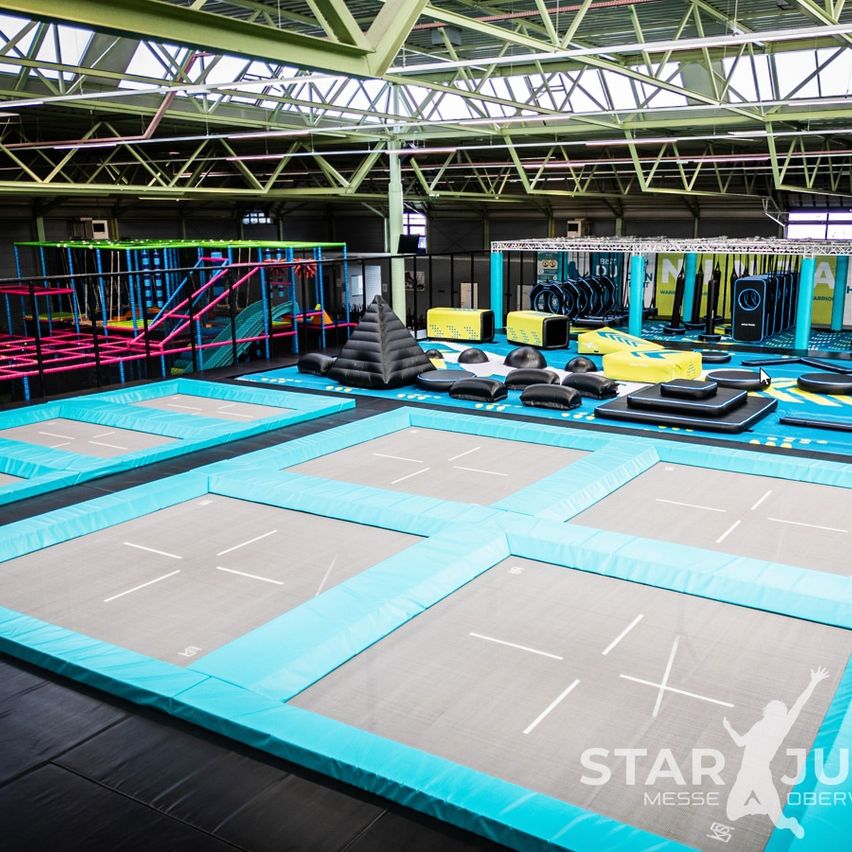 Ein Indoor-Trampolinpark mit mehreren blauen und grauen Trampolinen, einem pyramidenförmigen Hindernis und verschiedenen Trainingsgeräten. Eine Person springt auf den Trampolinen.