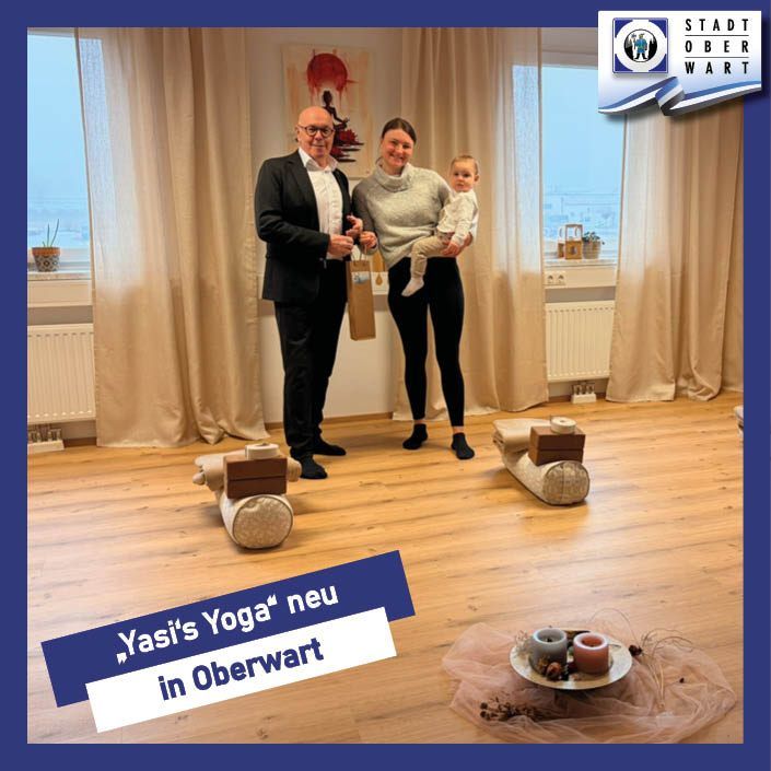 Ein Mann, eine Frau und ein Baby stehen in einem Raum mit Holzboden. Die Frau hält das Baby. Es gibt dekorative Baumstämme auf dem Boden und ein Banner mit deutschem Text.