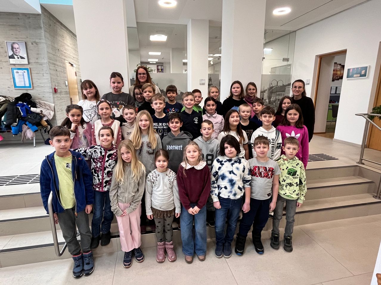 Eine Gruppe von Kindern und zwei Erwachsenen steht zusammen auf einer Treppe. Sie sind in lässiger Winterkleidung gekleidet. Die Umgebung ist drinnen, mit einem gefliesten Boden und einer Glaswand hinter ihnen.