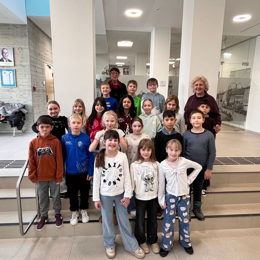 Eine Gruppe von Kindern und zwei Erwachsenen posiert für ein Foto auf einer Treppe in einem Gebäude. Sie tragen Freizeitkleidung, und der Hintergrund zeigt eine Glaswand und Bilderrahmen.