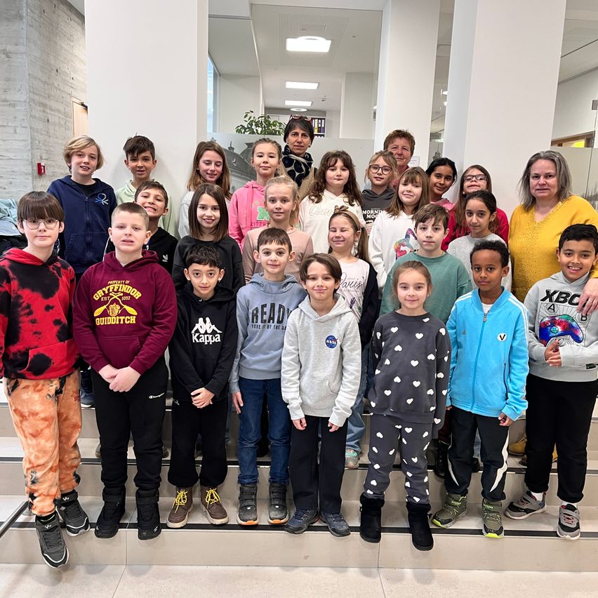 Eine Gruppe von Kindern, einige mit Brille, steht auf einer Treppe. Sie posieren für ein Foto mit einer lächelnden Frau. Die Kinder tragen verschiedene Jacken und Turnschuhe. Dahinter ist ein verschwommener Blick auf eine Wand und eine Pflanze zu sehen.