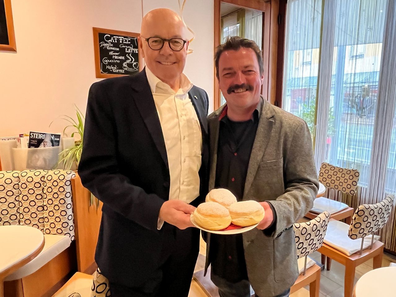 Zwei Männer in Anzügen halten eine Platte mit Donuts und lächeln. Dahinter steht ein Schwarzes Brett mit der Aufschrift 'Caffe'. Sie stehen in einem Raum mit Stühlen und einer Pflanze.