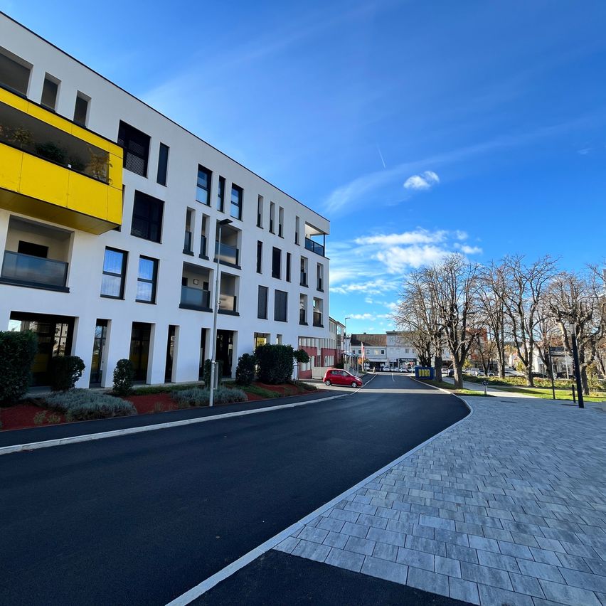 Eine leere Straße mit einem geparkten roten Auto, in der Nähe eines modernen Wohngebäudes. Die Straße hat einen gebogenen Bürgersteig mit Bäumen und Büschen an der Seite.