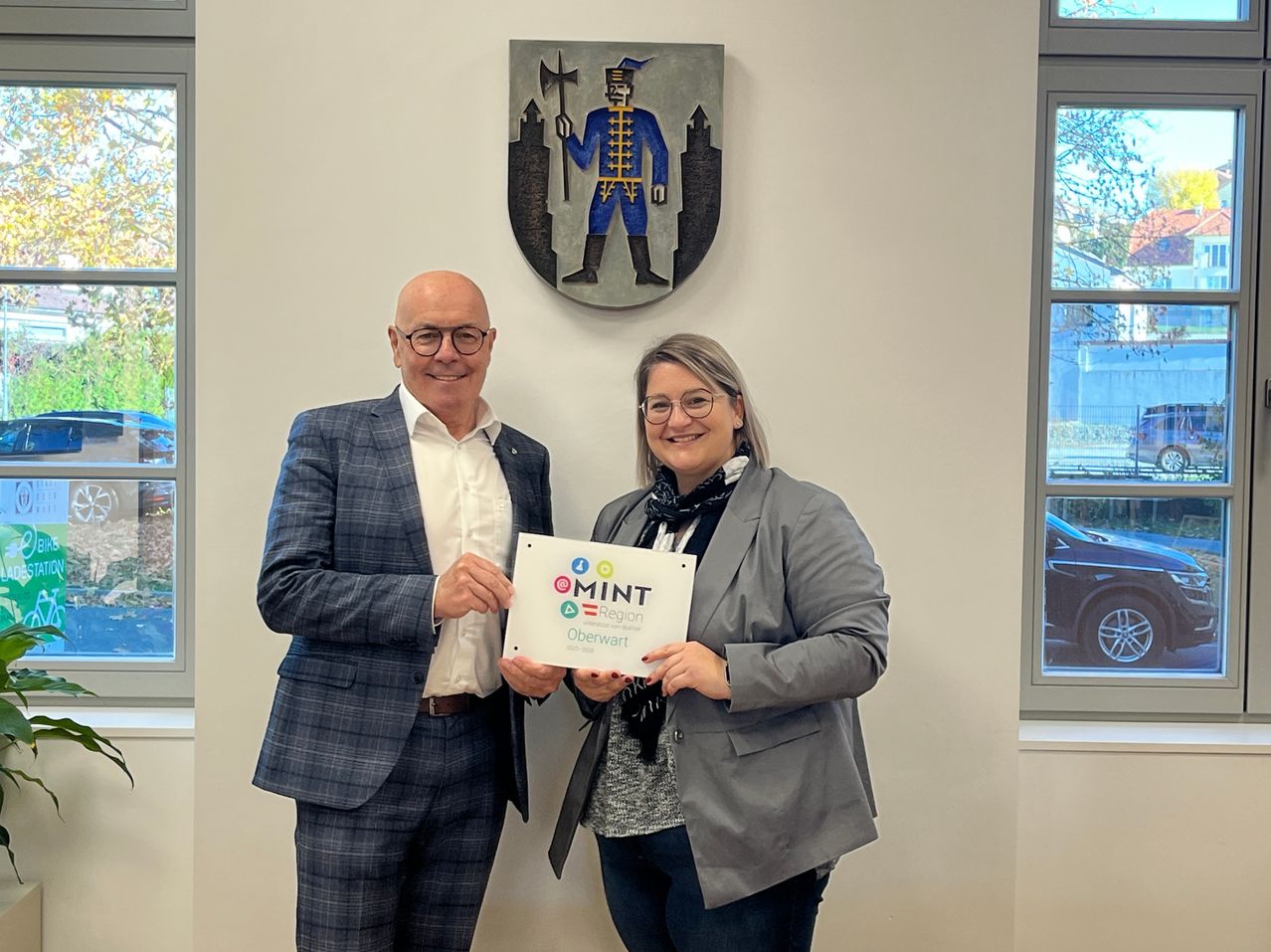 Ein Mann und eine Frau stehen in einem Raum und halten ein gerahmtes Zertifikat mit dem Text 'MINT Region Oberwart'. Dahinter befindet sich ein dekorativer Schild an der Wand.