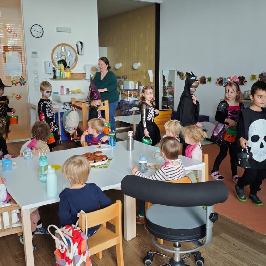 Eine Gruppe von Kindern in Halloween-Kostümen versammelt sich um einen Tisch mit Essen und Getränken. Eine Frau steht in der Nähe, möglicherweise eine Lehrerin oder Mitarbeiterin. Der Raum ist mit Halloween-Motiven dekoriert.