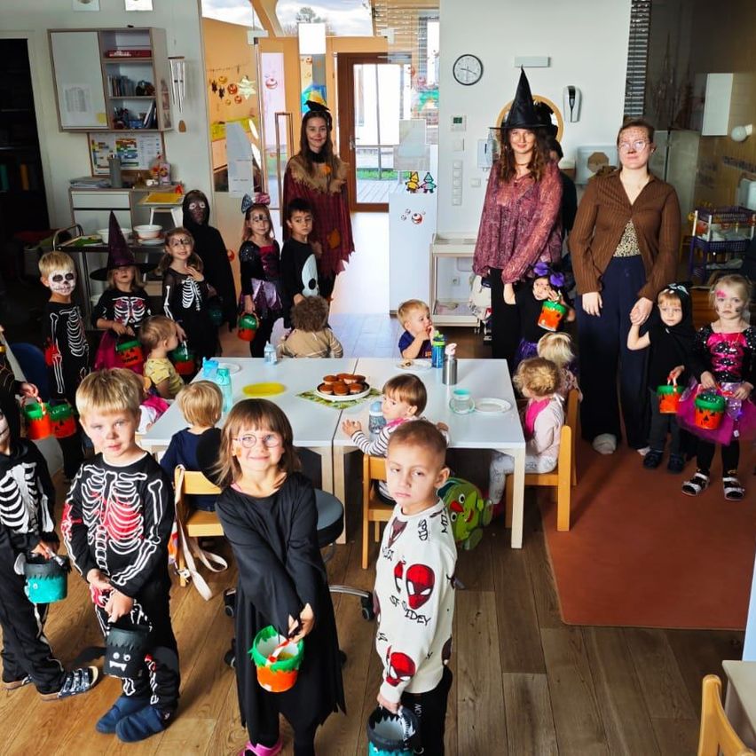 Eine Gruppe von Kindern in Halloween-Kostümen steht in einem Raum mit Erwachsenen, einer hält ein Baby. Ein Tisch ist mit Tellern, Tassen und Essen gedeckt. An der Wand hängt eine Uhr und ein Kühlschrank im Hintergrund.