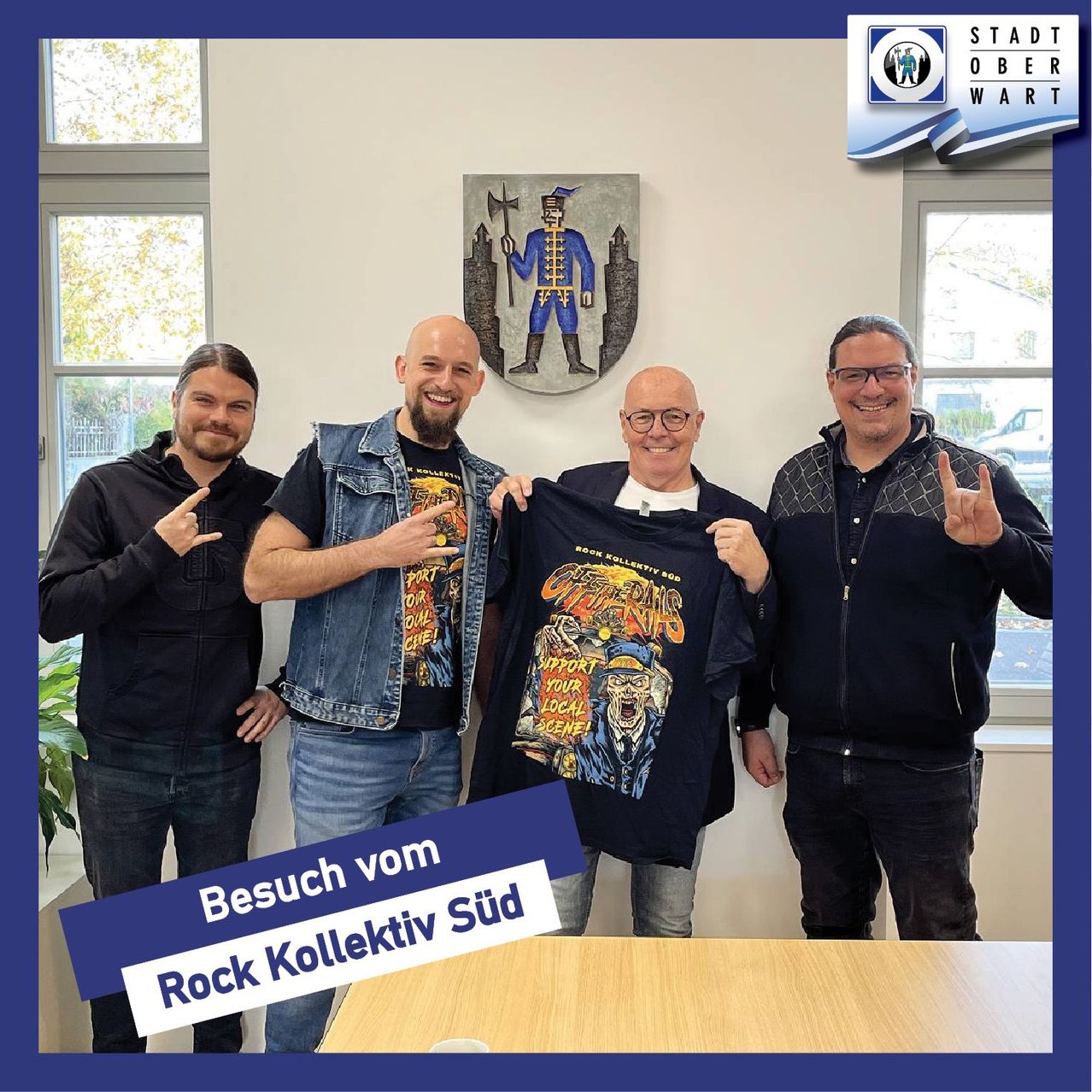 Vier Männer stehen in einem Raum, zwei mit Händen auf der Brust, einer hält ein schwarzes T-Shirt und einer zeigt ein Friedenszeichen. Hinter ihnen eine Wand mit einem deutschen Wappen und einem Fenster, das ein Auto zeigt.