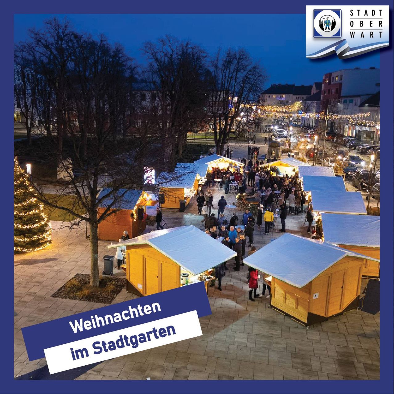 Luftaufnahme eines Weihnachtsmarktes mit Holzhütten, einem geschmückten Weihnachtsbaum und einer Menschenmenge unter dem Nachthimmel.