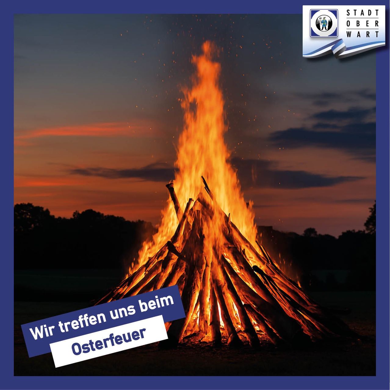 Ein großes Lagerfeuer brennt hell bei Dämmerung mit einem blauen Banner, das 'Wir treffen uns beim Osterfeuer' steht.