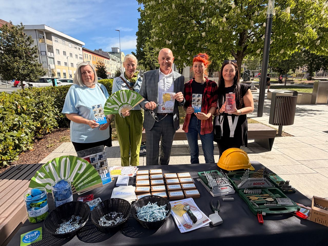 Five people stand behind a table filled with tools and brochures. They hold STOP signs and smile for the camera. Behind them, trees and a cityscape are visible.
