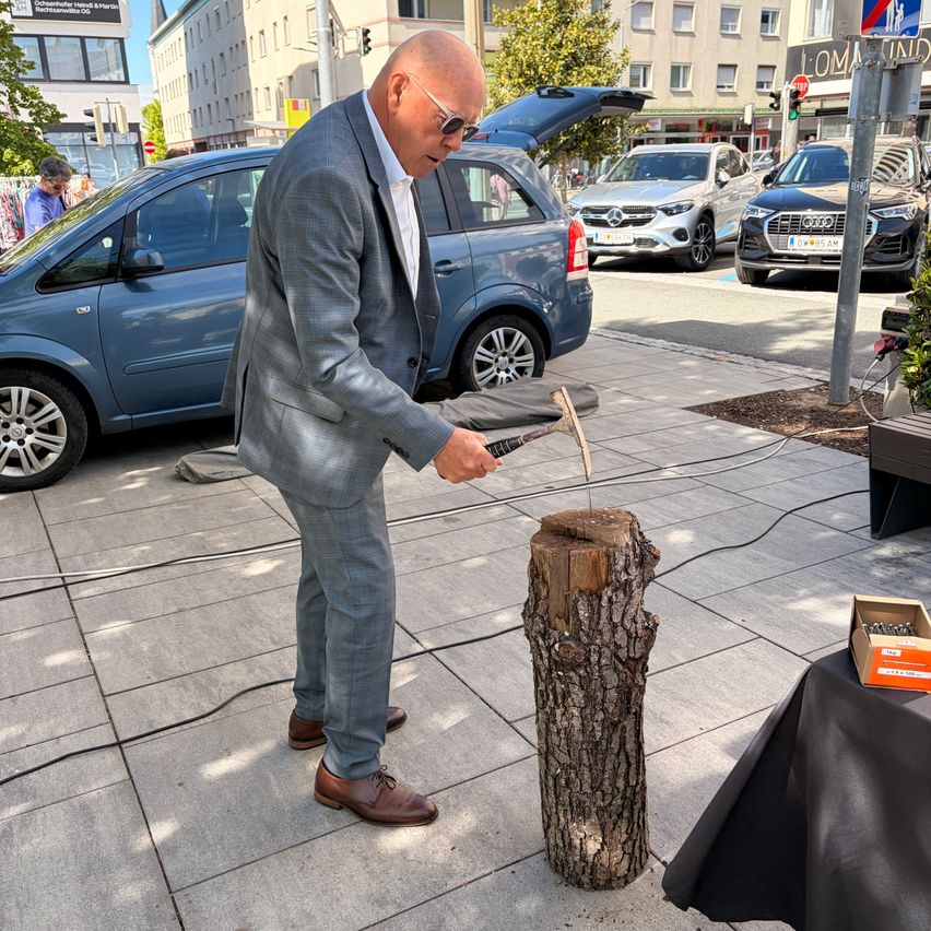 A man in a suit hammers a wooden post on a city sidewalk. Cars are parked nearby.