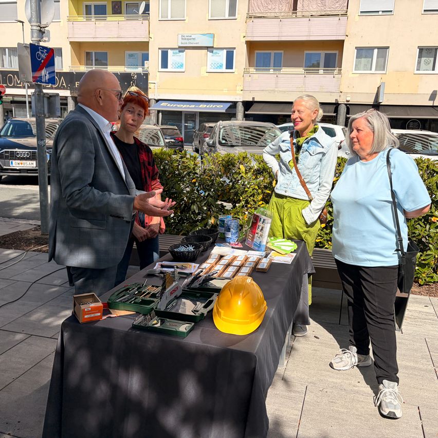 Four people stand by a table with tools and pamphlets in a city square. A man speaks to two women. Buildings and cars are in the background.