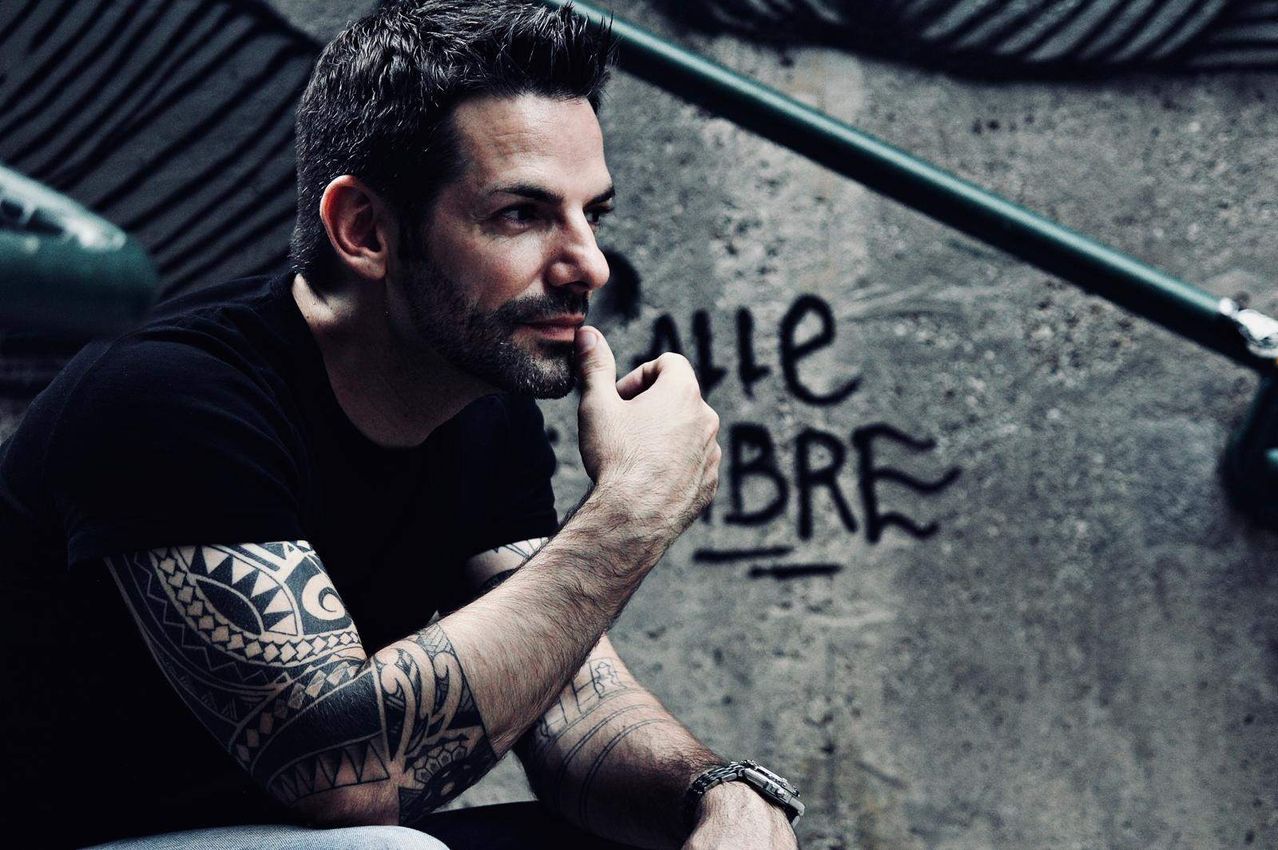 Ein Mann mit Tattoos an beiden Armen sitzt und schaut nach links, die Hand am Kinn. Hinter ihm ist eine Wand mit Graffiti.