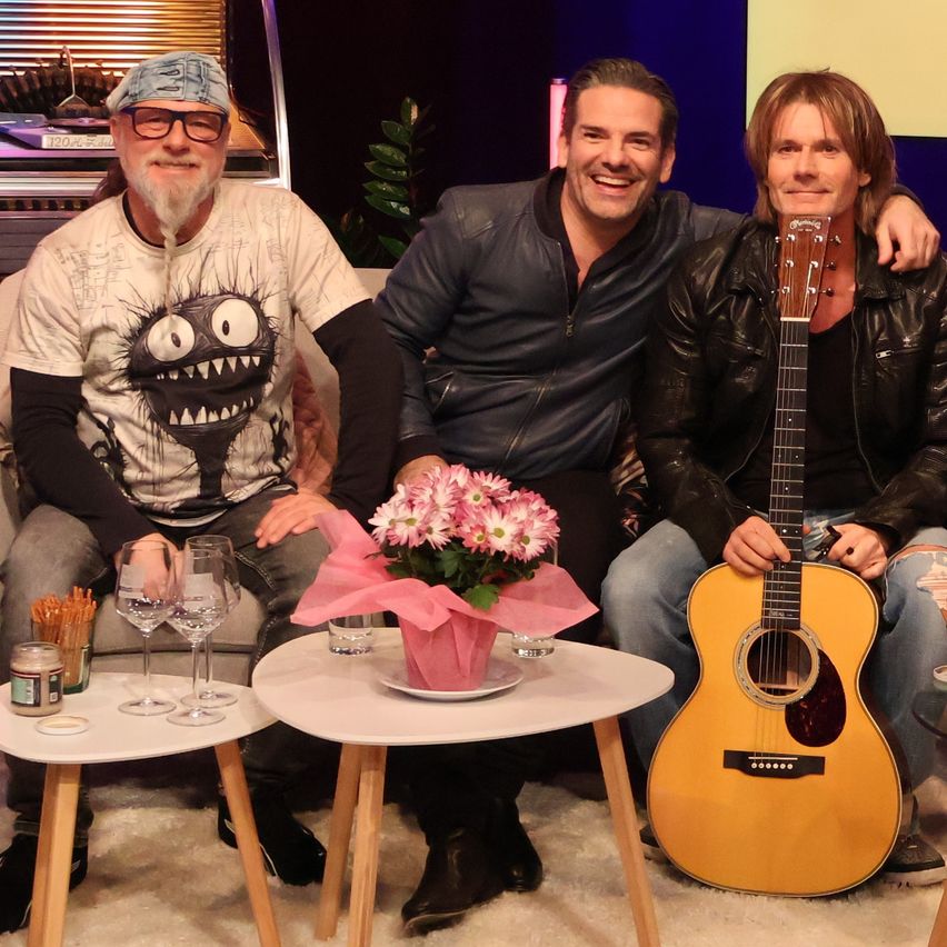 Drei Männer sitzen lächelnd zusammen auf einer Couch. Einer hält eine Gitarre, ein anderer hat einen Blumenstrauß und der dritte trägt einen Totenkopf. Sie sind von Beistelltischen mit Weingläsern und einem kleinen Glas umgeben.