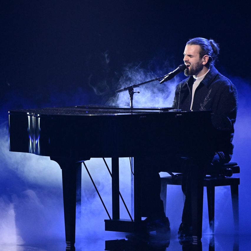 Ein Mann mit einem Dutt sitzt auf einem Hocker und spielt Klavier. Er singt in ein Mikrofon. Im Hintergrund ist Blau mit Raucheffekten zu sehen.