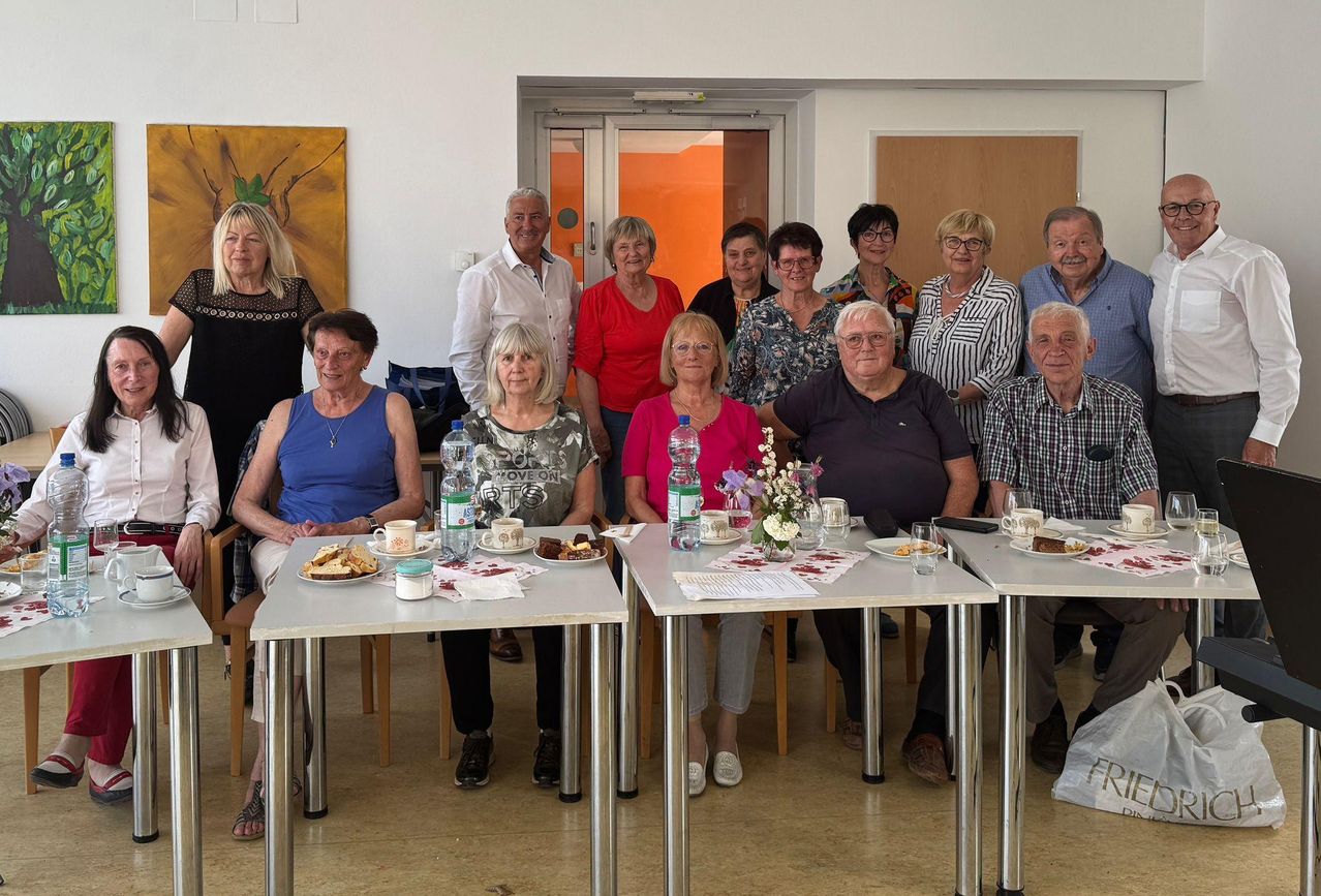 Eine Gruppe älterer Menschen sitzt um Tische in einem Raum, lächelt und posiert für ein Foto. Die Tische haben Blumen, Tassen und Teller. Der Raum hat weiße Wände, Türen und ein großes Gemälde.