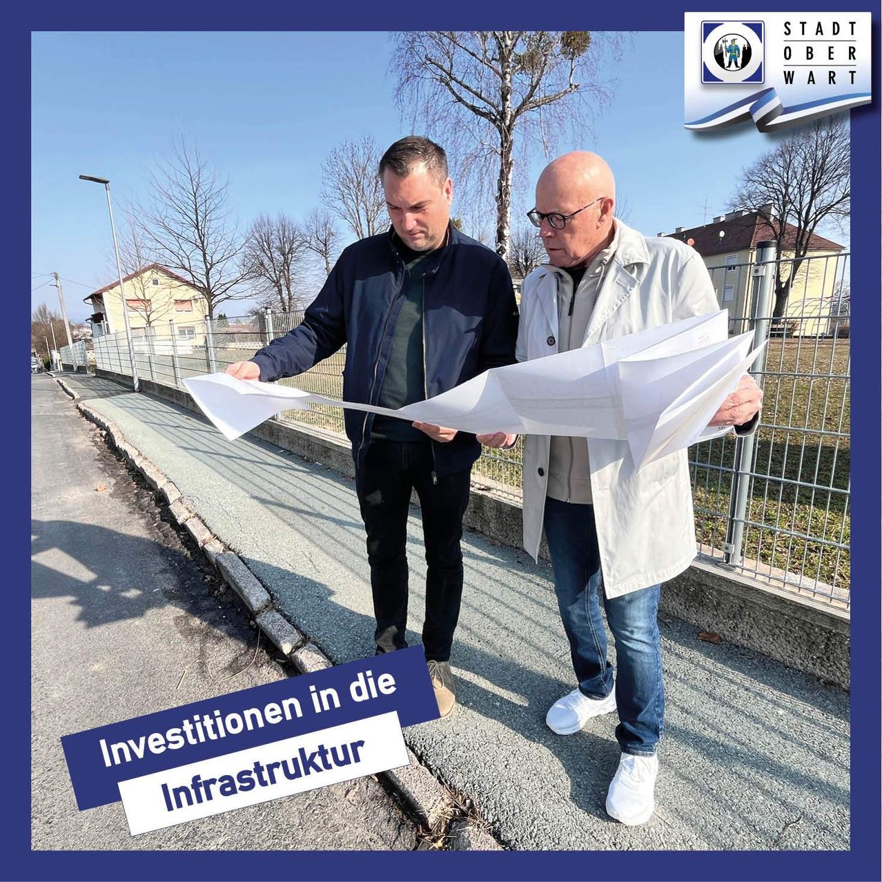 Zwei Männer stehen auf einem Bürgersteig, einer hält eine Karte in der Hand und der andere schaut sie an. Sie sind beide in Jacken gekleidet. Hinter ihnen steht ein Zaun mit einem Haus. Das Bild hat einen Wasserzeichen mit dem Text Stadt Oberwart.