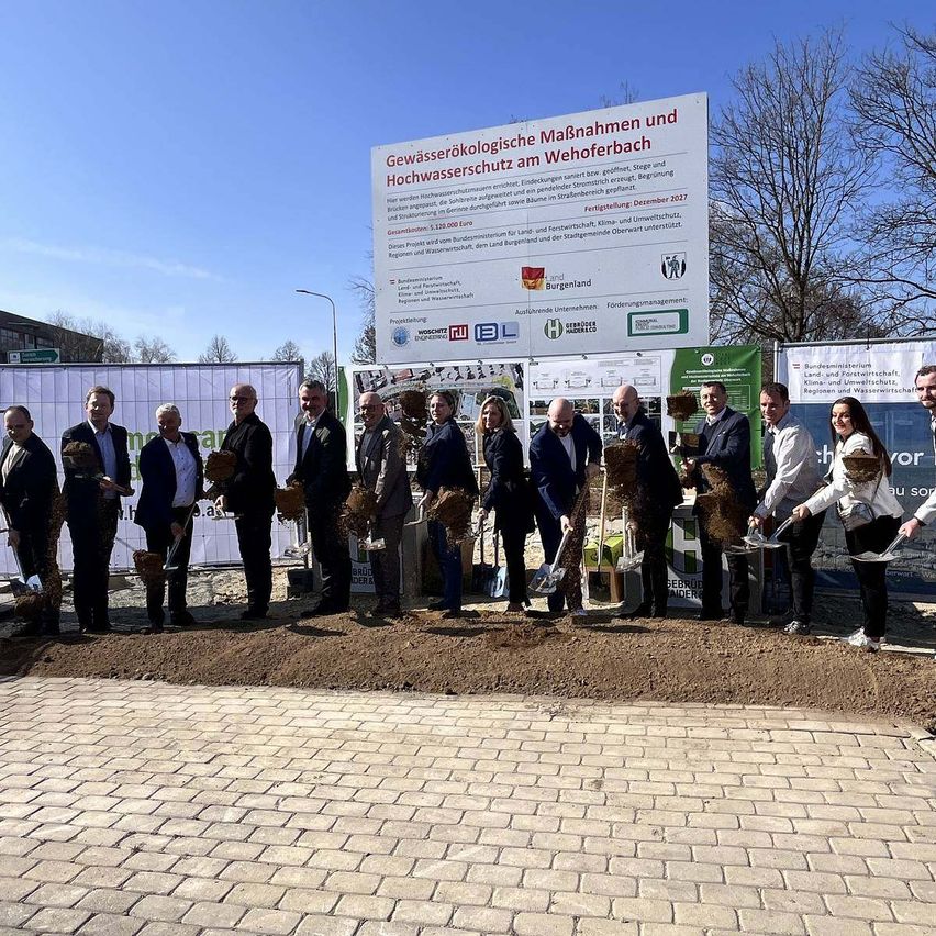 Eine Gruppe von Menschen in formeller Kleidung steht in einem mit Erde bedeckten Bereich mit Schaufeln und hält Erde in den Händen vor einem großen Schild.