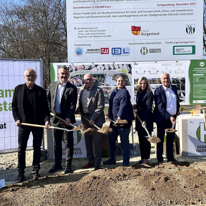 Eine Gruppe von Menschen in formeller Kleidung steht vor einem großen Banner und hält Schaufeln. Sie feiern wahrscheinlich eine Grundsteinlegungszeremonie. Das Banner zeigt Logos und Text im Zusammenhang mit dem Projekt, einschließlich Namen beteiligter Organisationen.