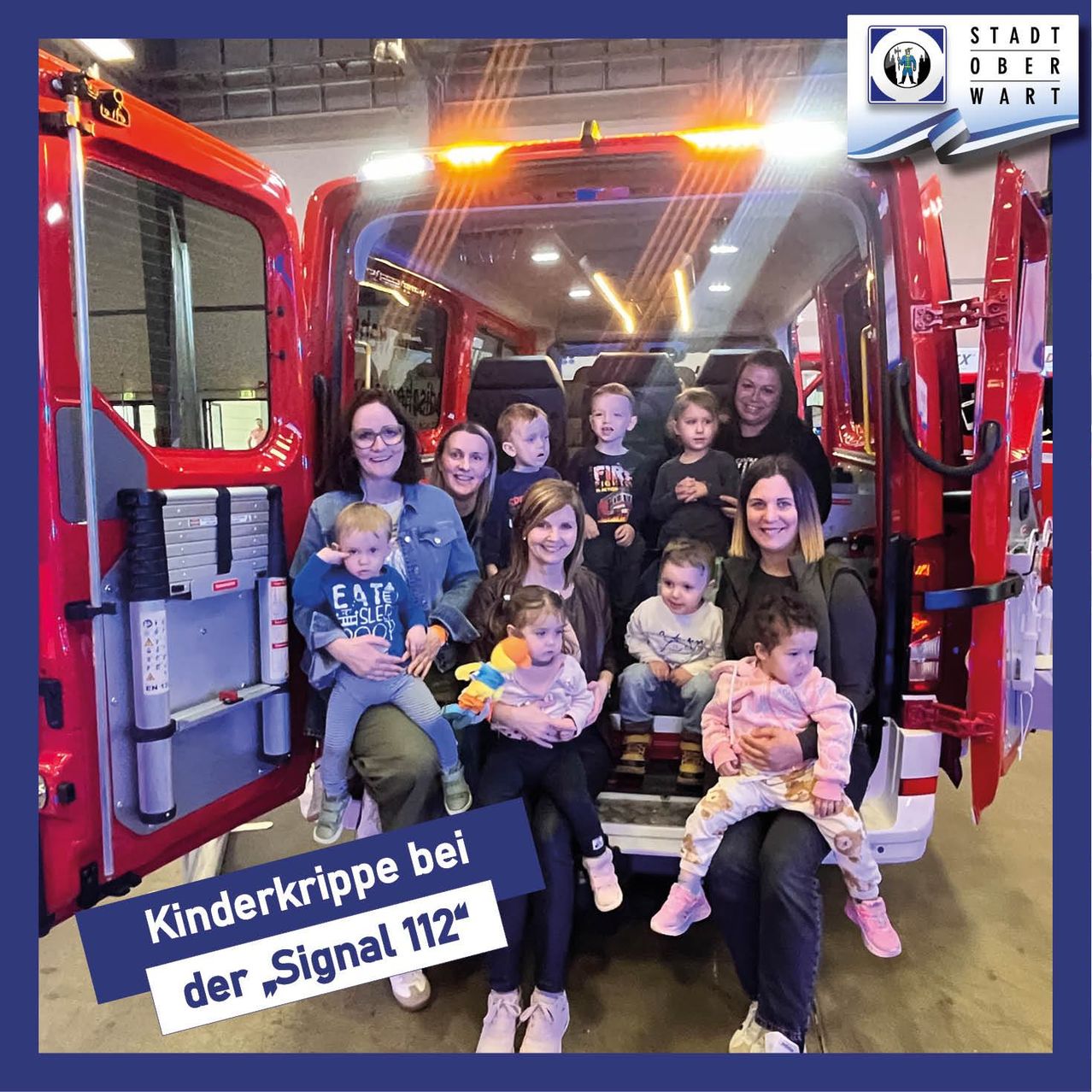 Eine Gruppe von Kindern und Erwachsenen posiert für ein Foto in einem roten Krankenwagen mit offenen Türen. Der Textüberlagerung lautet 'Kinderkrippe bei der Signal 112'.