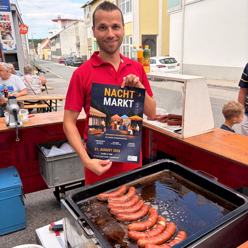 Ein Mann in einem roten Shirt hält ein Schild für Nacht Markt, mit Würsten, die in einer Pfanne vor ihm kochen.