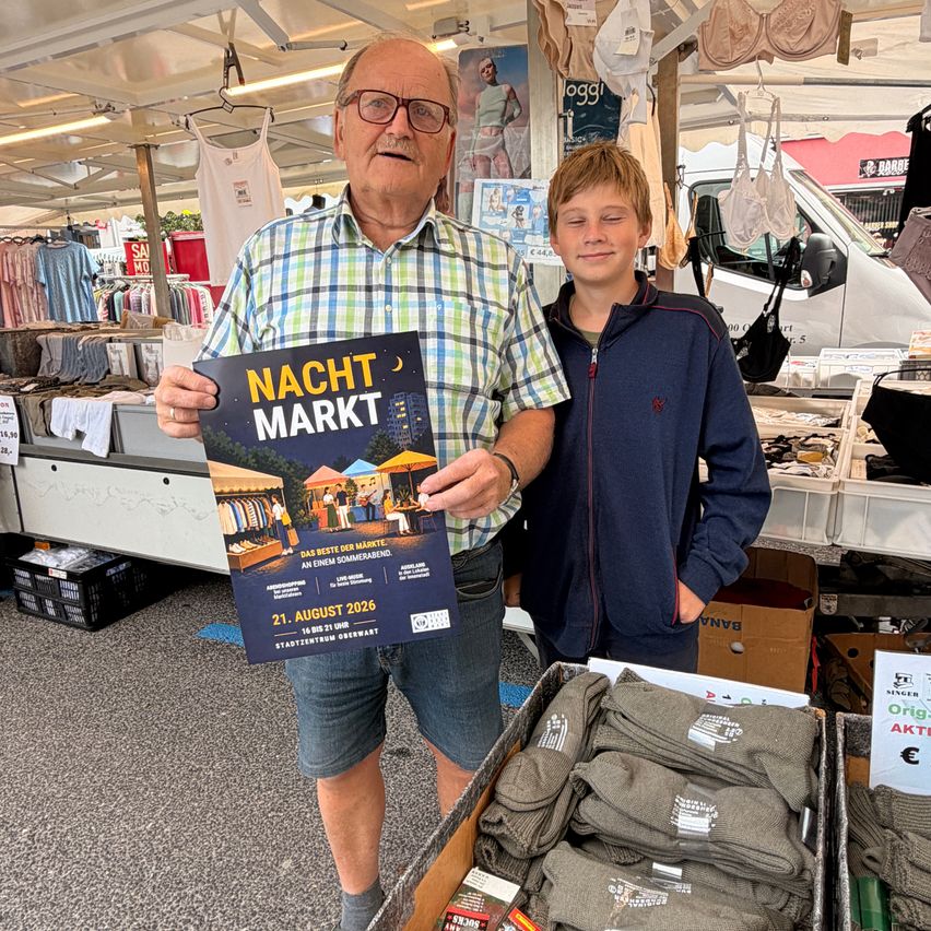 Ein älterer Mann und ein junger Junge stehen vor einem Kleidungsstand auf einem Markt und halten ein Schild für die Nacht Markt Veranstaltung am 21. August 2026.