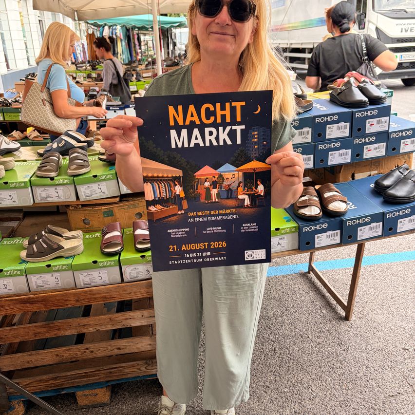 Eine Frau hält ein Plakat für Nacht Markt, eine Veranstaltung, die für den 21. August 2026 geplant ist, vor einem Schuhmarkt mit verschiedenen Schuhen im Angebot.
