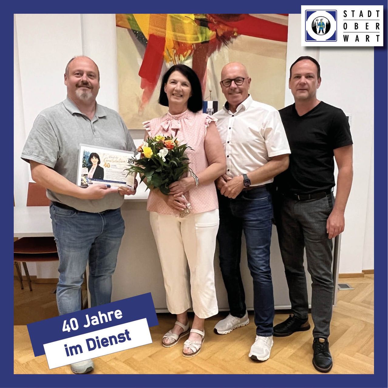 Vier Erwachsene stehen in einem Raum, lächeln und posieren für ein Foto. Eine Frau hält Blumen und ein Zertifikat. Der Mann auf der linken Seite hält ein Zertifikat und der Text lautet '40 Jahre im Dienst'. Ein Gemälde ist hinter ihnen.
