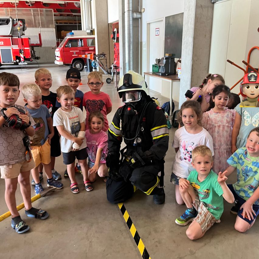 Eine Gruppe von Kindern steht in einer Feuerwache mit einem Feuerwehrmann. Der Feuerwehrmann kniet und ist von Kindern umgeben. Die Kinder lächeln und posieren für ein Foto.