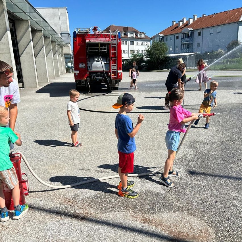 Kinder spielen mit einem Wasserschlauch eines Feuerwehrwagens unter Aufsicht von Erwachsenen an einem sonnigen Tag.