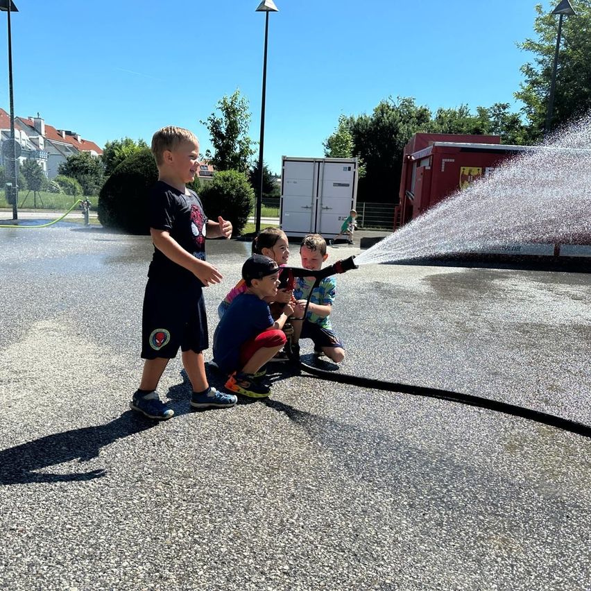 Vier Jungen sprühen Wasser aus einem Feuerwehrschlauch auf einem Parkplatz. Bäume und Gebäude sind im Hintergrund.