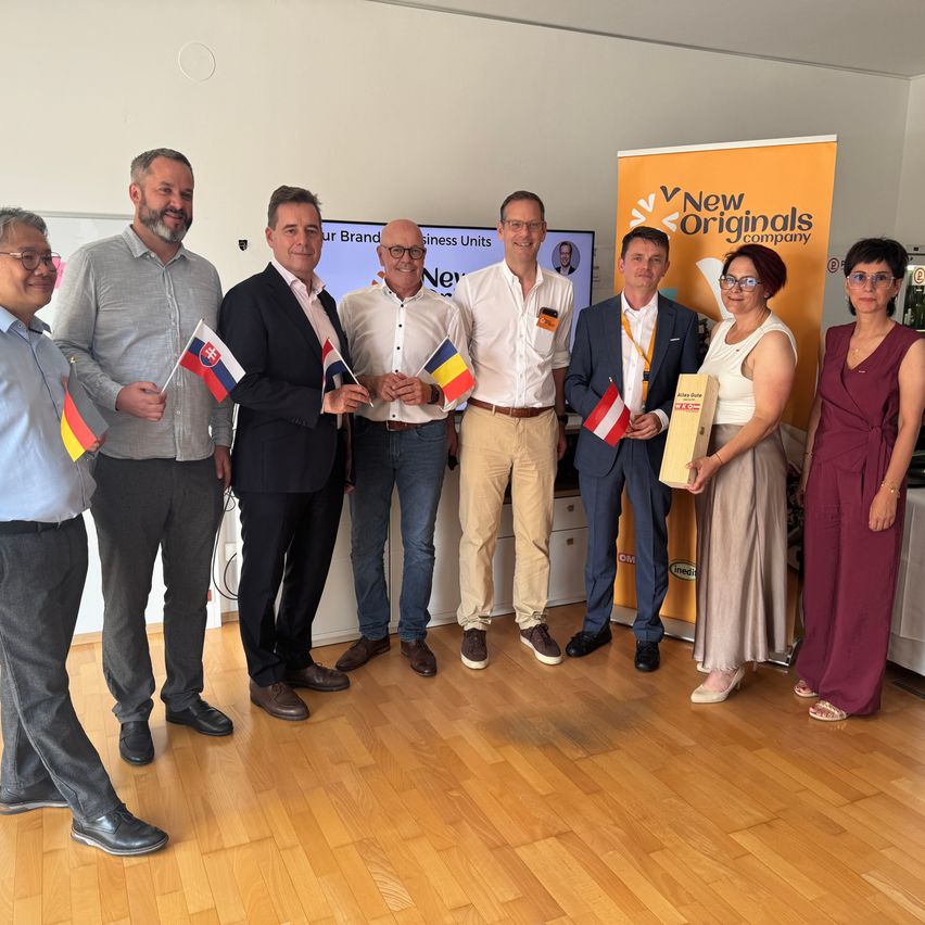 A group of individuals holding flags and an award stand on a wooden floor. A banner with New Originals is in the background.