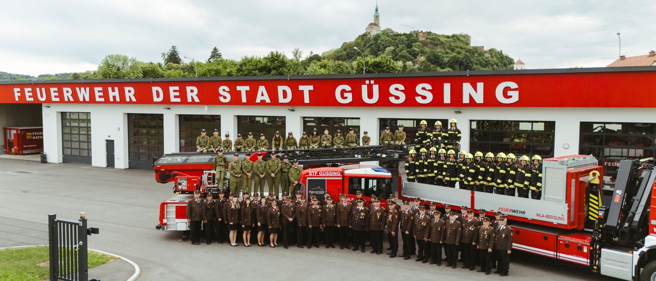 Eine Gruppe Feuerwehrleute in Uniform steht vor einem Feuerwehrauto, mit einem Gebäude, das mit 'Der Stadt Gussing' beschriftet ist, im Hintergrund.