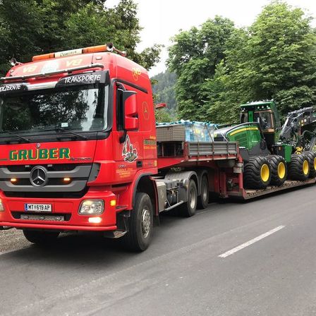 Ein großer roter LKW mit dem Wort Gruber auf der Front transportiert ein großes grünes Baufahrzeug auf einer Straße.