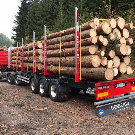 Ein großer Anhänger mit Baumstämmen, beschriftet mit Gruber Holz, geparkt auf einer Straße mit Bäumen im Hintergrund. Der Anhänger hat ein rotes und schwarzes Design und ist an einen roten LKW angeschlossen.