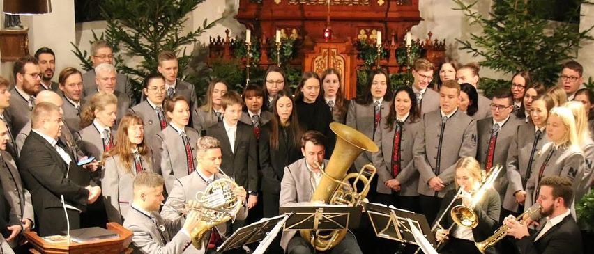 Ein großer Chor und ein Blechbläserensemble treten in einer Kirche auf, wobei einige Mitglieder auf der Bühne sitzen und andere stehen. Das Publikum sitzt vor ihnen, und einige Mitglieder halten ihre Instrumente.