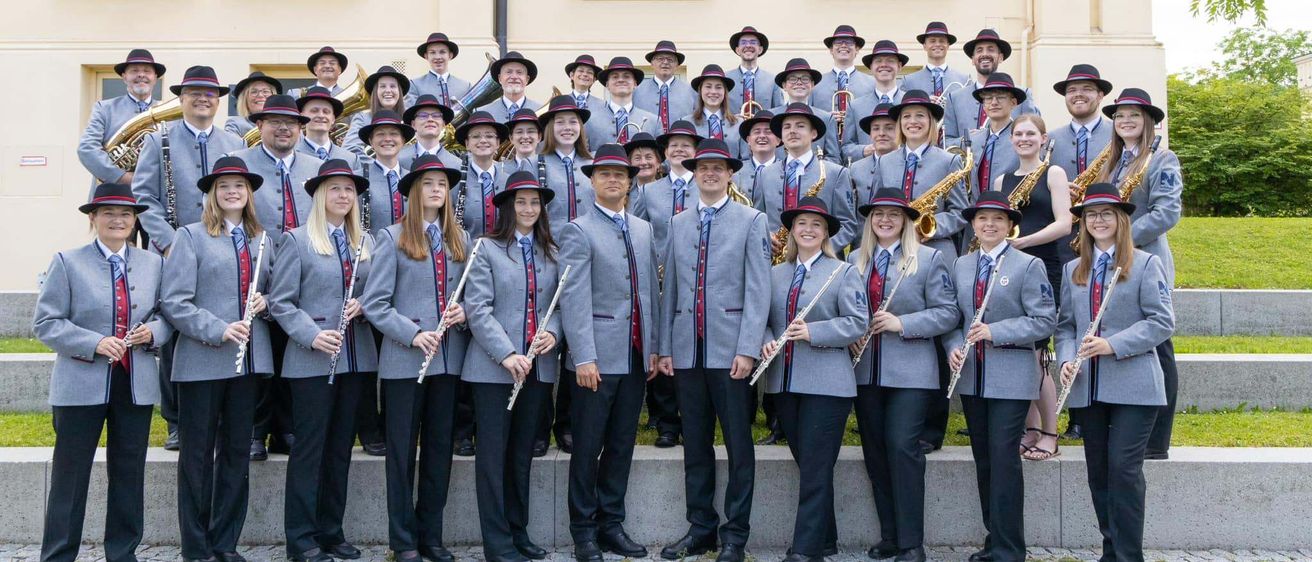 Eine Musikband in Uniform steht vor einem Gebäude. Sie halten verschiedene Musikinstrumente wie Flöten, Saxophone und Trompeten. Sie tragen Hüte und Anzüge mit roten und weißen Schärpen.