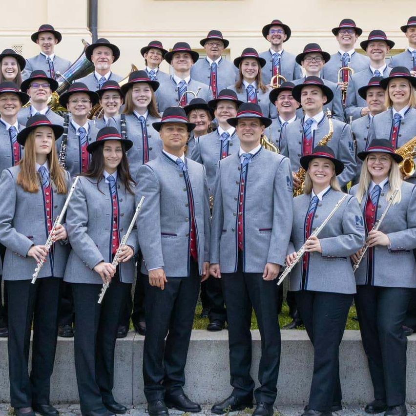 Eine Musikband in Uniform steht vor einem Gebäude. Sie halten verschiedene Musikinstrumente wie Flöten, Saxophone und Trompeten. Sie tragen Hüte und Anzüge mit roten und weißen Schärpen.