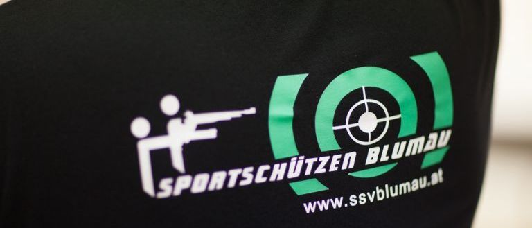Ein schwarzes T-Shirt zeigt ein weißes Logo einer Person, die mit einem Gewehr schießt, und einen grünen Kreis mit einem Zielsymbol. Der Text lautet SPORTSCHUTZEN BLUMAU und www.ssvblumau.at.