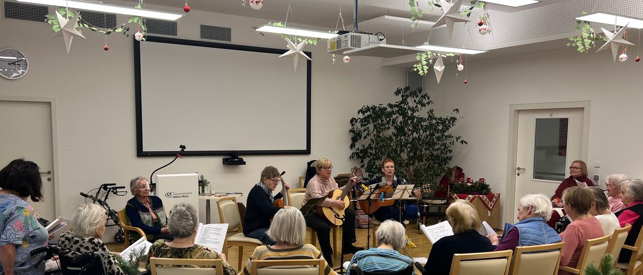 Eine Gruppe älterer Frauen sitzt in Stühlen in einem Raum, einige spielen Gitarre. Eine Leinwand ist an der Wand montiert, und Weihnachtsdekorationen schmücken die Decke.