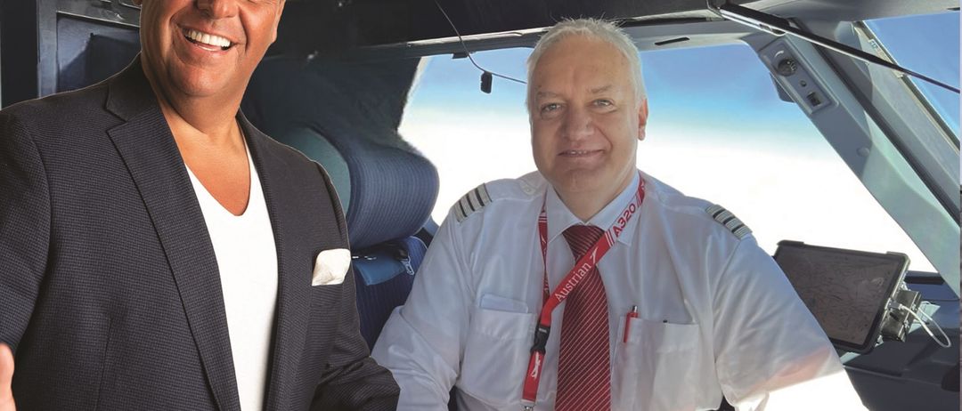 Zwei Männer sitzen in der Cockpit eines Flugzeugs. Der Mann auf der linken Seite lächelt und trägt einen Anzug. Der Mann auf der rechten Seite trägt ein weißes Hemd und eine rote Krawatte.