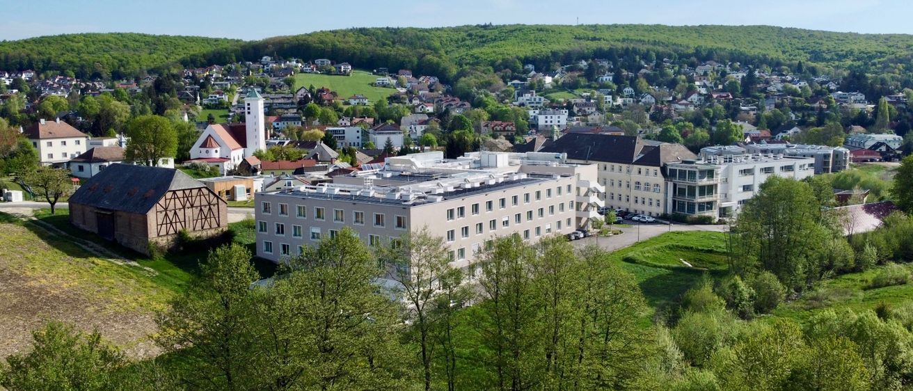 An aerial view of a large building with many windows surrounded by trees, a grassy area, and a hill in the distance.
